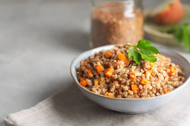 Photo of Boiled buckwheat cereal with carrot, parsley and butter in a bowl on a gray background. Traditional Russian dish on gray background with copy space