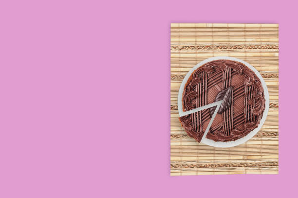 istock One cuted chocolate cake with cream and leaf on white ceramic round plate and bamboo rug on pink table in kitchen. Top view. Copy space 1146249326