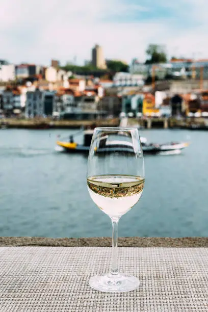 Selective focus of white wine glass overlooking Vila Nova de Gaia embankment at Cais da Ribeira on the River Douro in Porto, Portugal - reflection of bridge on glass