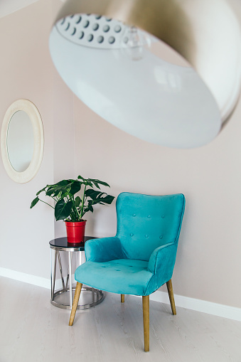 Conceptual light  interior of living room with blue armchair, metallic standing lamp,  glass round coffee table , oval mirror on white wall, monstera plant in red vase on the floor. Concept of minimalizm.