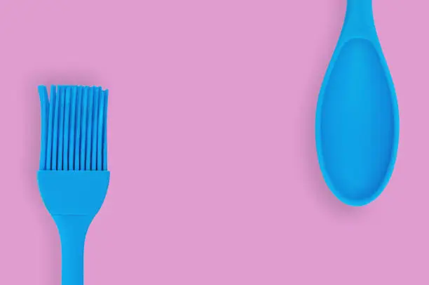 Blue rubber or silicone brush and spoon with plastic handles for confectionery on pink table in kitchen. Flat lay. Copy space for your text