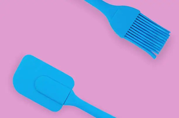 Blue rubber or silicone brush and spatula with plastic handles for confectionery on pink table in kitchen. Flat lay. Copy space for your text