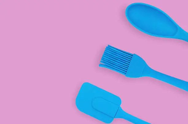 Blue rubber or silicone brush, spoon and spatula with plastic handles for confectionery on pink table in kitchen. Flat lay. Copy space for your text