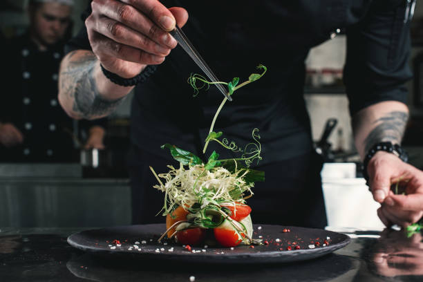 küchenchef veredelt gesunden salat auf einem schwarzen teller mit pinzette. fast bereit, es auf einem tisch zu servieren - speisen und getränke stock-fotos und bilder