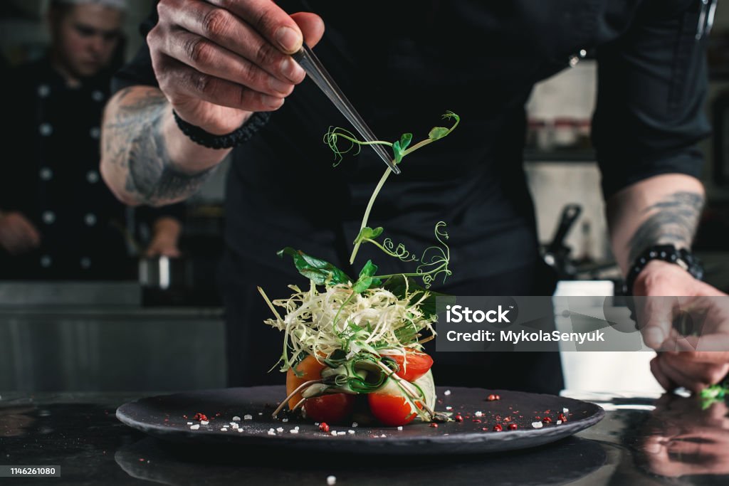 Chef terminando una ensalada sana en un plato negro con pinzas. casi listo para servirlo en una mesa - Foto de stock de Chef libre de derechos