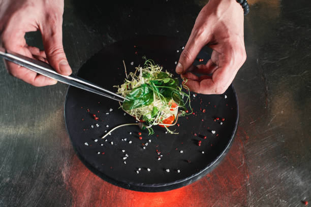 Chef finishing healthy salad on a black plate with tweezers. almost ready to serve it on a table Chef finishing healthy salad on a black plate with tweezers. almost ready to serve it on a table. indoors restaurant hotel work tool stock pictures, royalty-free photos & images