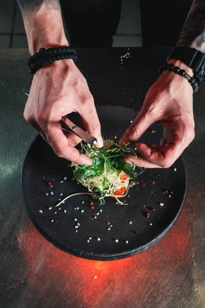 Chef finishing healthy salad on a black plate with tweezers. almost ready to serve it on a table Chef finishing healthy salad on a black plate with tweezers. almost ready to serve it on a table. indoors restaurant hotel work tool stock pictures, royalty-free photos & images