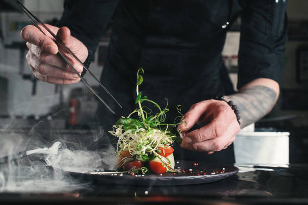 küchenchef veredelt gesunden salat auf einem schwarzen teller mit pinzette. fast bereit, es auf einem tisch zu servieren - feinschmecker essen stock-fotos und bilder