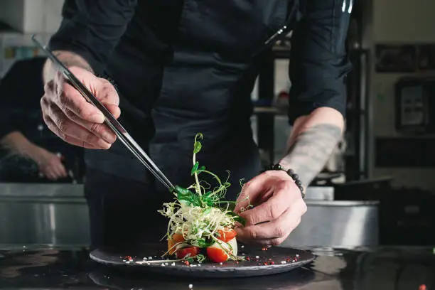 Chef finishing healthy salad on a black plate with tweezers. almost ready to serve it on a table.