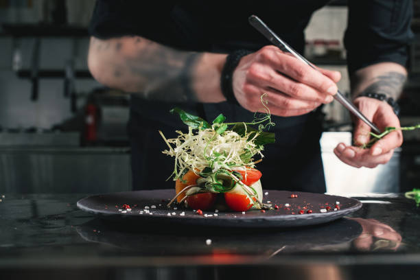 Chef finishing healthy salad on a black plate with tweezers. almost ready to serve it on a table Chef finishing healthy salad on a black plate with tweezers. almost ready to serve it on a table. indoors restaurant hotel work tool stock pictures, royalty-free photos & images