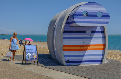 Eastbourne, Sussex, UK - August 1, 2018: Lady in blue sun dress reading information about the rentable stripy beach hut called spyglass,  on the beach in Eastbourne.