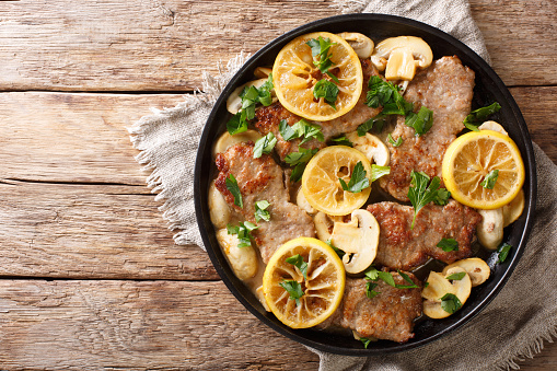 Tasty Italian fillet of veal scaloppini with mushrooms and lemons in sauce close-up in a frying pan on the table. Horizontal top view from above
