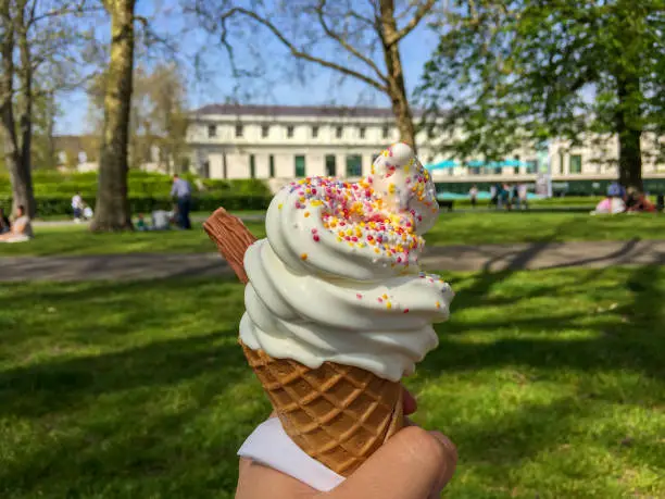 Person Holding Ice-Cream Cone With Chocolate Flake & Sprinkles In Summer Sunshine In a Public Park