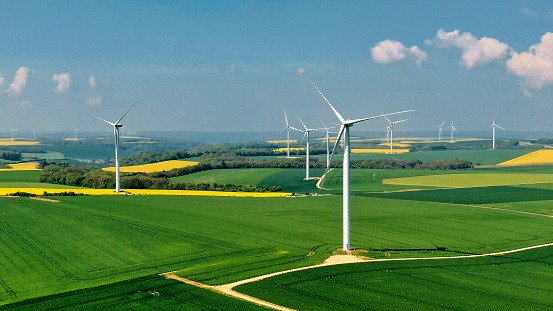 French landscape with a wind farm above rapeseed fields and forests under an azure blue sky dotted with a few small white clouds.