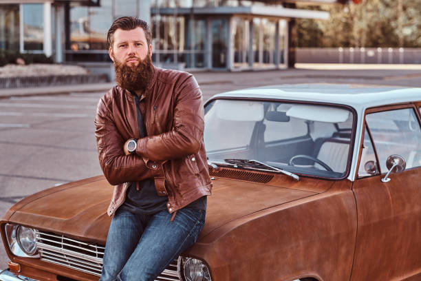 istock Bearded male dressed in brown leather jacket posing with crossed arms while leaning on tuned retro car in the city parking. 1146290766