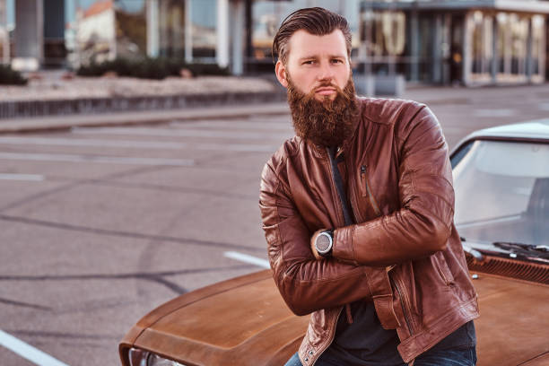 istock Bearded male dressed in brown leather jacket posing with crossed arms while leaning on tuned retro car in the city parking. 1146291359