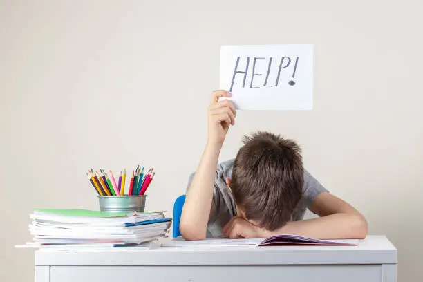 Photo of Sad tired frustrated boy sitting at the table with many books and holding paper with word Help. Learning difficulties, education concept.
