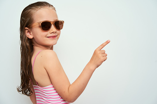 Smiling little preschool girl with wet hair photographed against white background wearing swimsuit and sunglasses pointing up with finger towards empty space