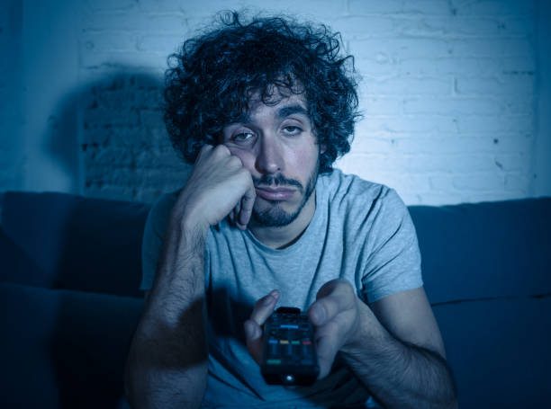 Young bored man on couch using TV remote control zapping for another movie or show late at night. Looking disinterested and sleepless. In entertainment People insomnia and Sedentary lifestyle concept. stock photo