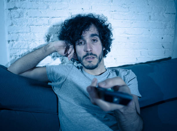 istock Young bored man on couch using TV remote control zapping for another movie or show late at night. Looking disinterested and sleepless. In entertainment People insomnia and Sedentary lifestyle concept. 1146320119