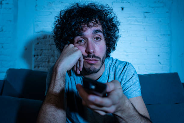 joven aburrido hombre en el sofá usando el control remoto de tv zapping para otra película o espectáculo tarde en la noche. buscando desinteresado y sin dormir. en entretenimiento personas insomnio y concepto de estilo de vida sedentario. - television boredom men sofa fotografías e imágenes de stock