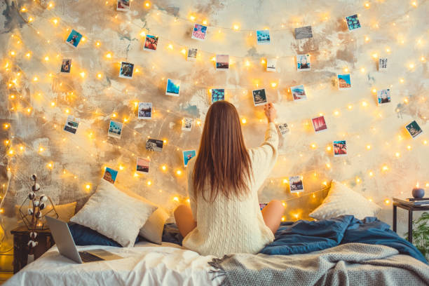 Young woman weekend at home decorated bedroom nostalgia Young woman creative weekend at home sitting looking at the photos on lighted wall memories nostalgia surrounding wall photos stock pictures, royalty-free photos & images