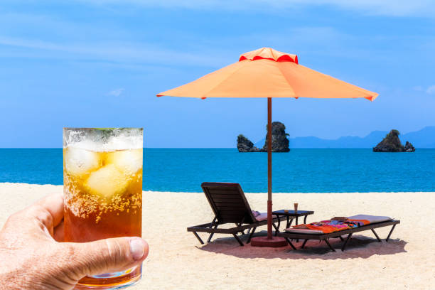 Hand holding glass of rum and coke with ice against sunloungers and a parasol on an empty Langkawi beach, Malaysia