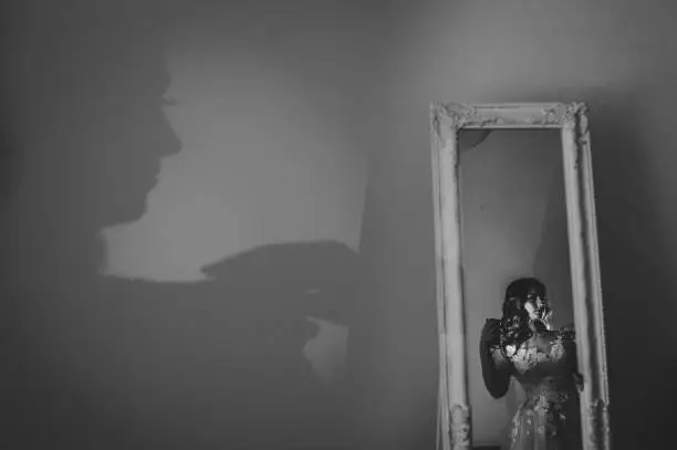 Photo of Artwork, noise, grain. Shadow of a woman. Portrait young attractive bride in dress stand in front of the mirror. Reflection body contour in the mirror on room. Rear view. Black and white photo.