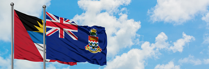 Antigua and Barbuda with Cayman Islands flag waving in the wind against white cloudy blue sky together. Diplomacy concept, international relations.