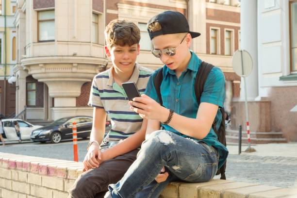 retrato al aire libre de dos niños de 13, 14 años de edad, hablando en la calle de la ciudad, amigos riendo, mirando el teléfono móvil - 13 14 years fotografías e imágenes de stock
