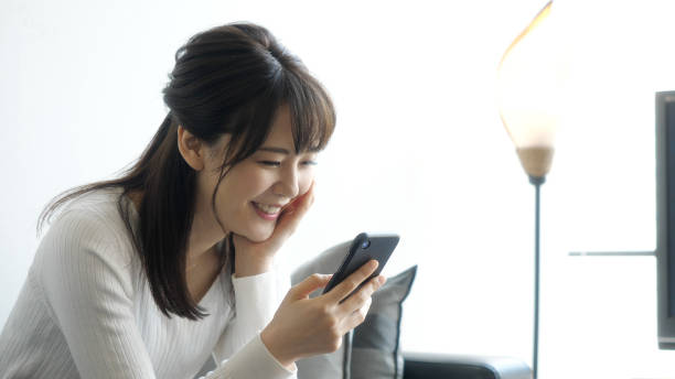 リビングルームでスマートフォンを使用して、魅力的なアジアの女性。 - 目線 ストックフォトと画像