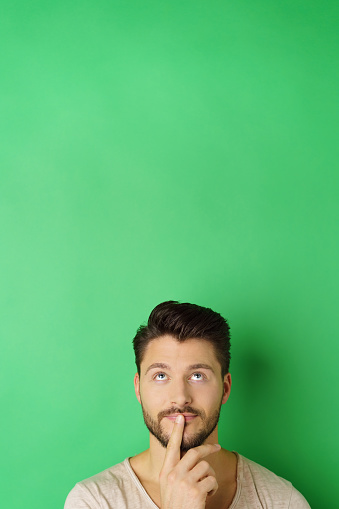 Portrait of young thoughtful bearded man looking up against green background with copy space