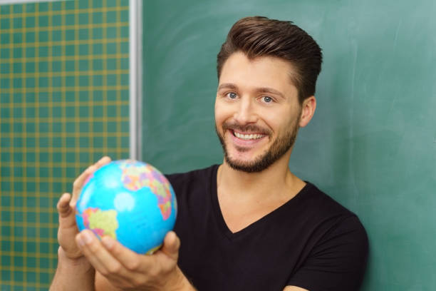 Geography teacher holding up a world globe in his hands with a happy smile in front of a chalkboard