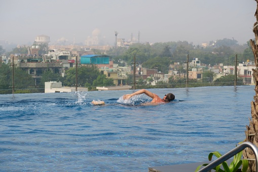 Stock photo showing a caucasian man doing the front crawl in an infinity swimming pool with a view overlooking the Taj Mahal in the distance. The pool has been surfaced with a series of matching blue and turquoise square blue mosaic tiles.