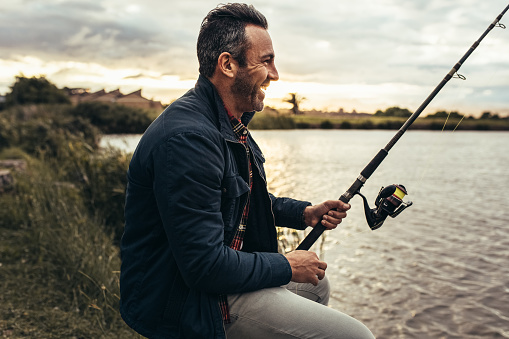 Smiling man sitting beside a lake and fishing. Side view of a happy man sitting near a lake holding a fishing rod.