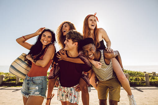 Five young people enjoying outdoors on their summer holiday. Group of multi-ethnic men and women enjoying piggyback ride on summer day.