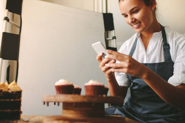 Taking pictures of pastries Young woman taking a pictures while cooking tasty pastry in the kitchen. Happy woman taking a picture of cupcakes on a wooden board with mobile phone while standing at the kitchen. confectioner photos stock pictures, royalty-free photos & images