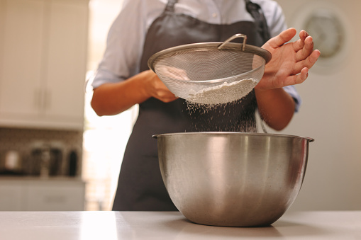 Close up of female pastry chef sifting flour in a bowl to make dough in kitchen. Woman baker baking in home kitchen.