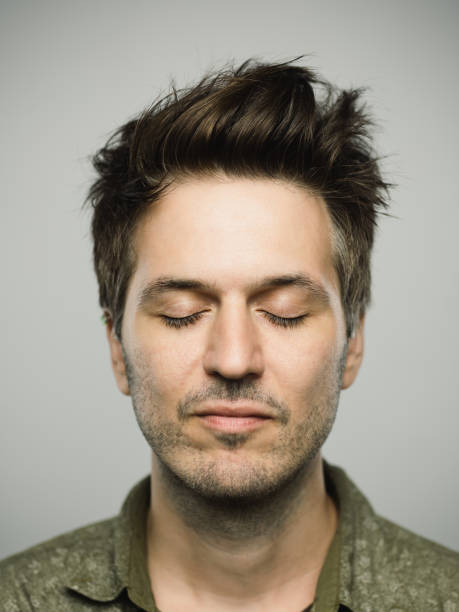 Real caucasian man with blank expression and eyes closed Close up portrait of young adult man with blank expression and eyes closed against gray white background. Vertical shot of caucasian real people resting in studio with brown hair and modern spiky haircut. Photography from a DSLR camera. Sharp focus on eyes. eyes closed stock pictures, royalty-free photos & images