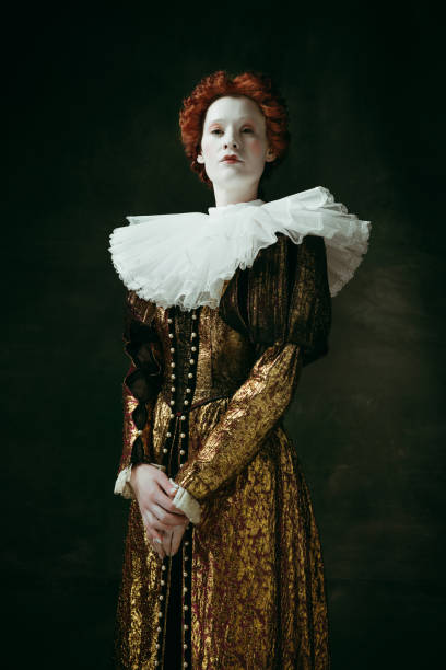 Medieval young woman as a duchess Attention and serious. Medieval redhead young woman in golden vintage clothing as a duchess standing crossing hands on dark green background. Concept of comparison of eras, modernity and renaissance. duchess photos stock pictures, royalty-free photos & images