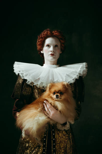 Medieval young woman as a duchess My best friend. Medieval redhead young woman in golden vintage clothing as a duchess holding puppy and standing on dark green background. Concept of comparison of eras, modernity and renaissance. queen royal person photos stock pictures, royalty-free photos & images