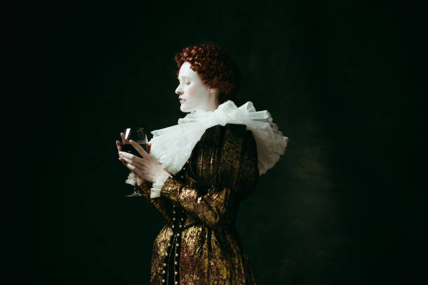 Medieval young woman as a duchess One part of me. Medieval redhead young woman in golden vintage clothing as a duchess holding a glass with red wine on dark green background. Concept of comparison of eras, modernity and renaissance. duchess photos stock pictures, royalty-free photos & images