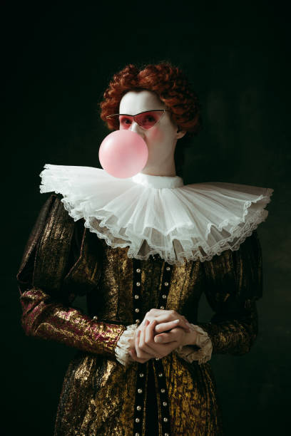 Medieval young woman as a duchess Medieval redhead young woman in golden vintage clothing as a duchess with red sunglasses blowing a bubblegum on dark green background. Concept of comparison of eras, modernity and renaissance. dark pink dress stock pictures, royalty-free photos & images