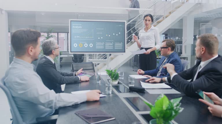 In the Corporate Meeting Room: Female Analyst Uses Digital Interactive Whiteboard for Presentation to a Board of Executives, Lawyers, Investors they Applaud. Screen Shows Company Growth Data with Animated Graphs