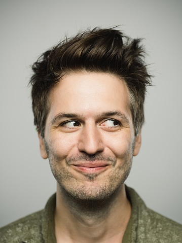 Close up portrait of young adult man with happy expression looking to the side against gray white background. Vertical shot of caucasian real people smiling and observing in studio with brown hair and modern spiky haircut. Photography from a DSLR camera. Sharp focus on eyes.
