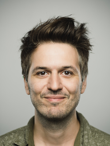 Close up portrait of young adult man with happy expression looking at camera against gray white background. Vertical shot of caucasian real people smiling in studio with brown hair and modern spiky haircut. Photography from a DSLR camera. Sharp focus on eyes.