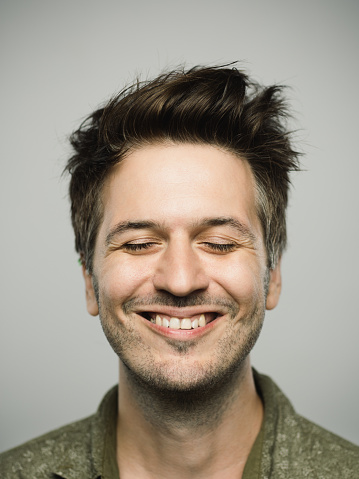 Close up portrait of young adult man with excited expression and eyes closed against gray white background. Vertical shot of caucasian real people relaxed in studio with brown hair and modern spiky haircut. Photography from a DSLR camera. Sharp focus on eyes.