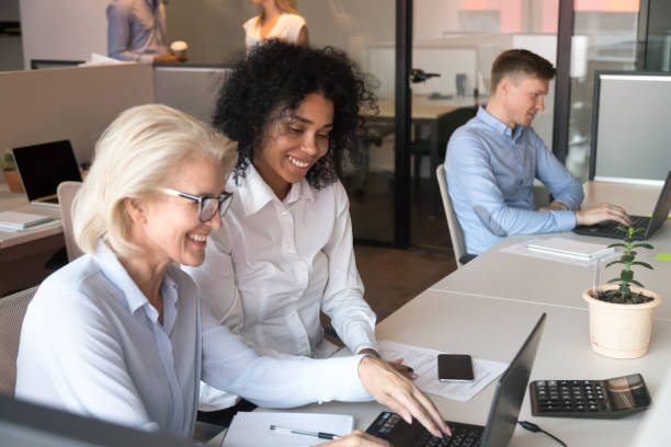 lächelnde alte mentorin, die afrikanischer praktikanten mit laptop im büro hilft - leadership business women senior adult stock-fotos und bilder