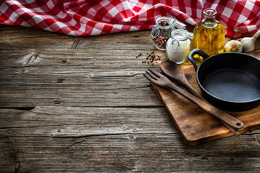 Cooking backgrounds: high angle view of a black cast iron pan, salt, olive oil, peppercorns arranged at the right of a rustic wooden kitchen table leaving a useful copy space for text and/or logo at the center left. A gingham tablecloth is at the top of the table making a frame. Predominant colors are brown and red. Low key DSRL indoors photo taken with Canon EOS 5D Mk II and Canon EF 24-105mm f/4L IS USM Wide Angle Zoom Lens