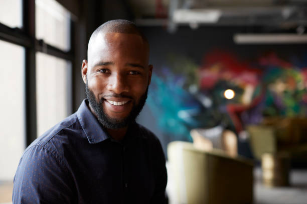 medio adulto negro masculino creativo en un área social de la oficina que se acerca a la cámara sonriendo, de cerca - cabeza afeitada fotografías e imágenes de stock